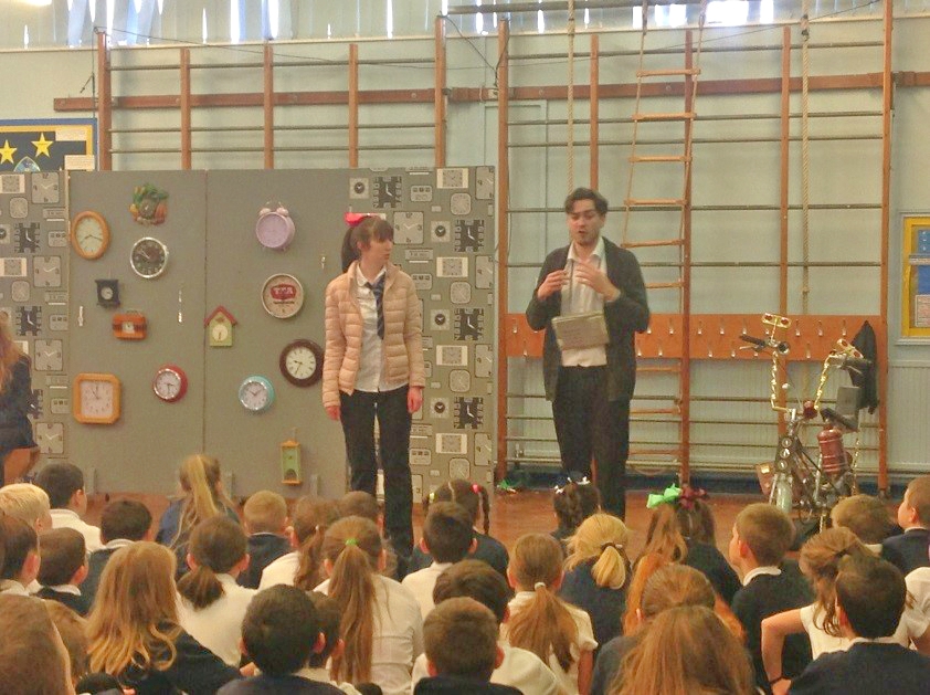 Two teachers are speaking to a group of children sat on the floor in the school hall.