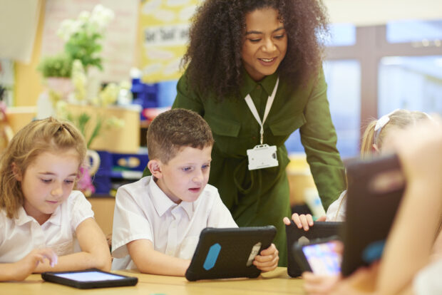 A teacher stands over a group of school children whilst they use tablets.