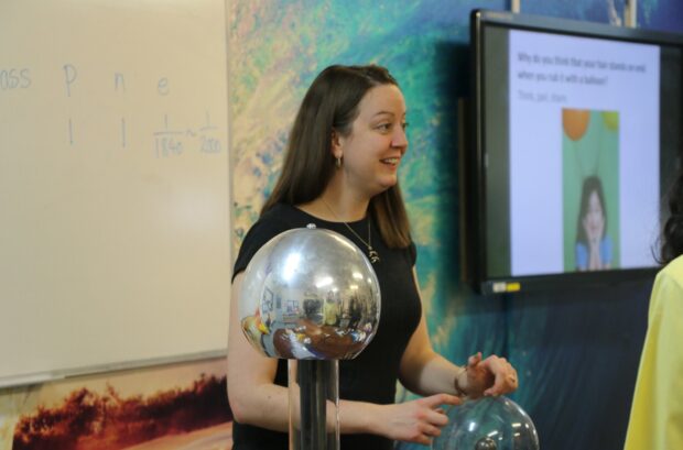 Emma Russo, physics teacher standing at the front of the classroom leading a class using a silver physics object