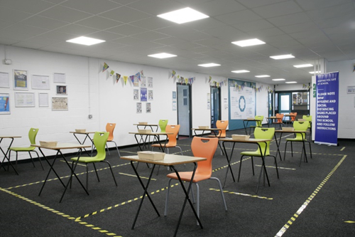 A large school room with single desks spaced evenly apart with yellow and black tape marking the boundaries in on the floor.