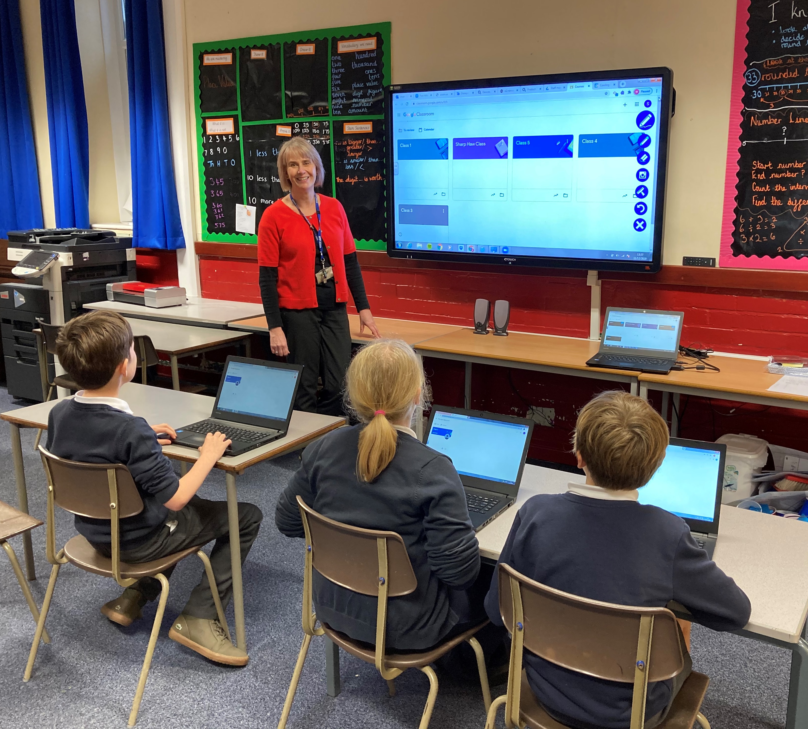 A teacher at the front of a classroom smiles at the camera, three primary pupils sit at desks with laptops