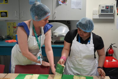 A member of staff standing with a student in a catering class. They are wearing aprons and hair nets. The student is holding ingredients.