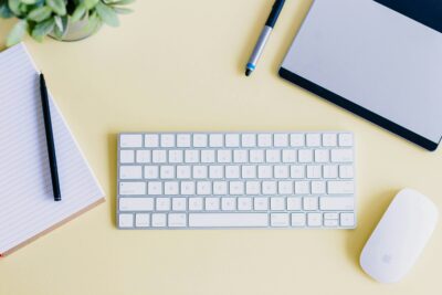 Laptop, keyboard, notepad and pen, and mouse on yellow surface