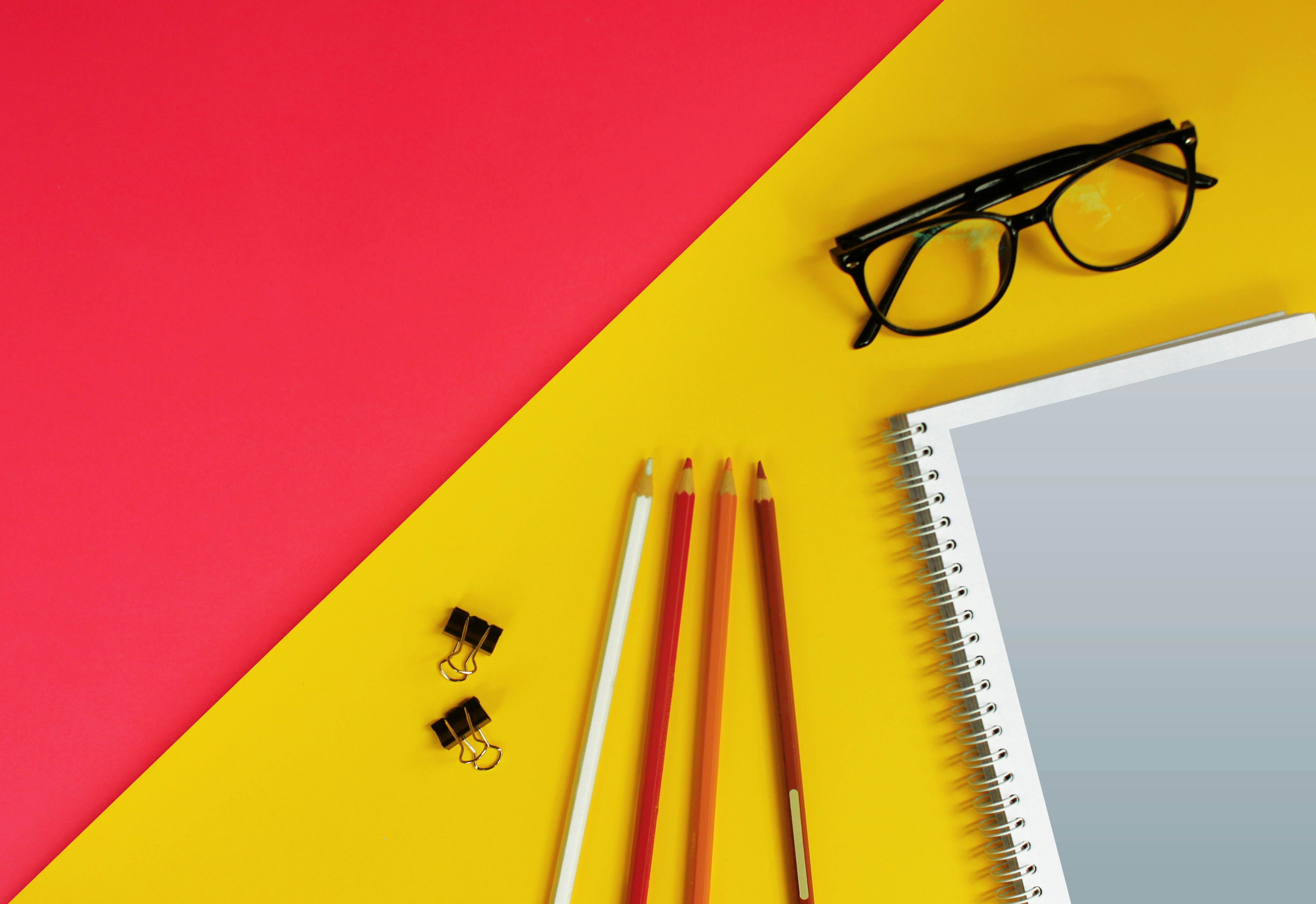 A pair of glasses, clips, pencils, and a book on a yellow and pink background