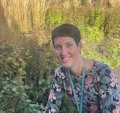 A teacher smiling looking at the camera, surrounded by a green garden with tall plants.